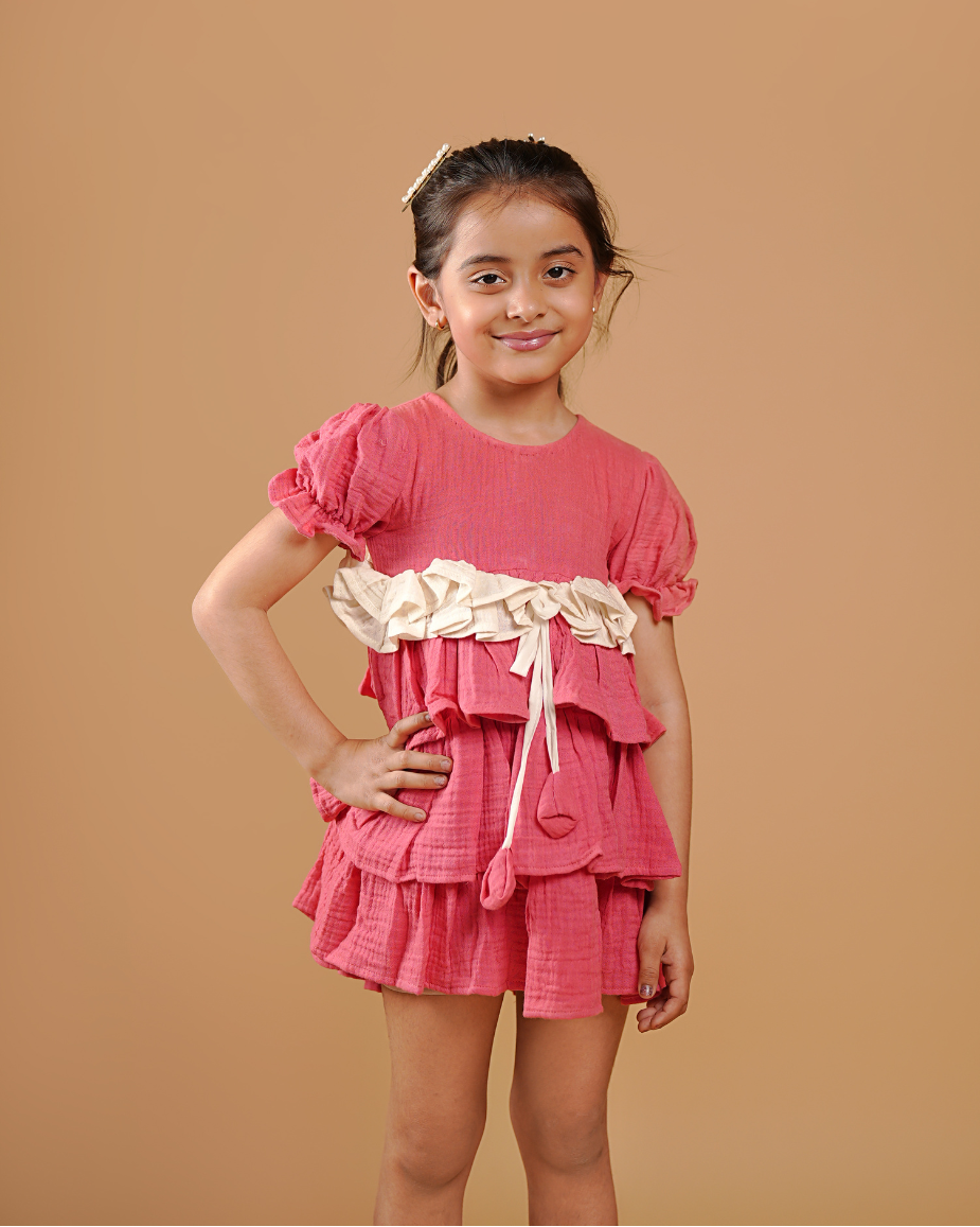 Young girl wearing a pink dress with ruffles and a belt against a brown background