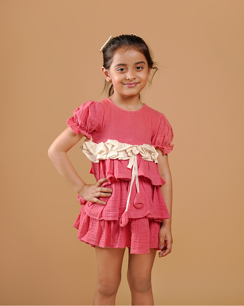Young girl wearing a pink dress with ruffles and a belt against a brown background
