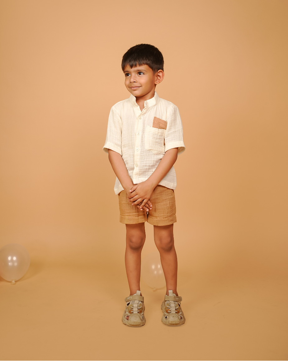 Young boy standing against a beige background wearing a white shirt and brown shorts.