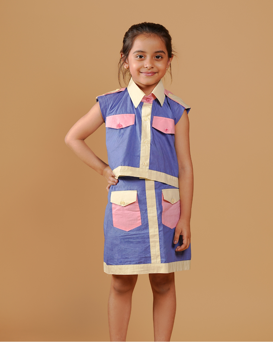 Young girl wearing a blue dress with pink pockets against a brown background