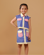 Young girl wearing a blue dress with pink pockets against a brown background