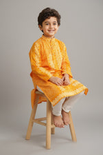 Baby Kids Cloths boy wearing a vibrant orangish-yellow patterned kurta with white pyjamas sitting on a wooden stool.